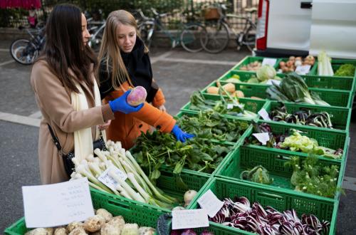 Twee vrouwen bekijken groenten van een groentekraam. 