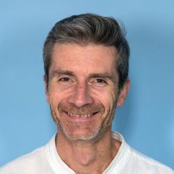 Een portretfoto van een witte man met kort haar en een baard. Hij draagt een witte t-shirt.